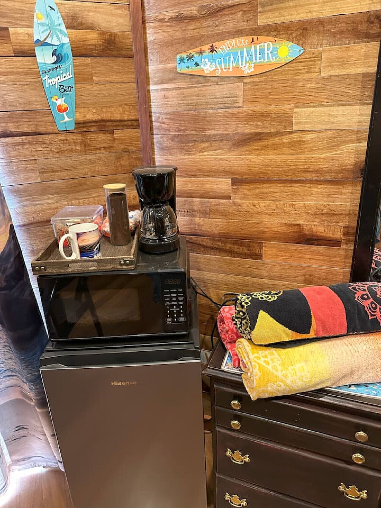 Ett litet kök med mikrovågsugn, kaffebryggare och förvaringsbehållare. Det finns en väggpanel av trä med en skylt som visar ordet ”Sommar” och en surfbräda som dekoration.
