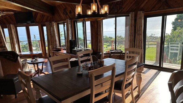 A dining area with a wooden table and chairs, large windows with a view of the ocean, and a chandelier. - Magic Skies Surfs Up Room (Hilo)
