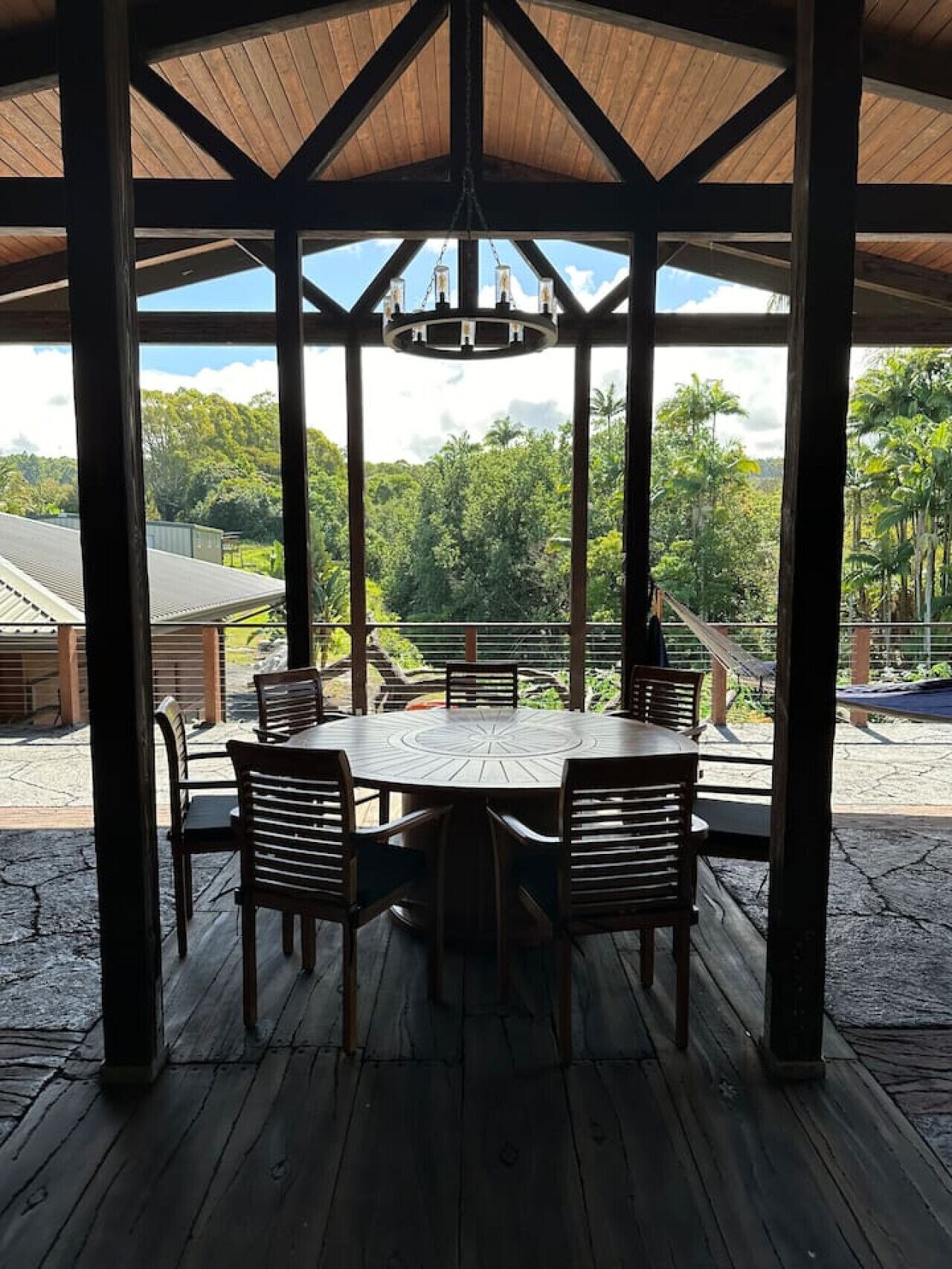 A covered outdoor dining area with a wooden table and chairs, large windows, and a view of lush greenery.