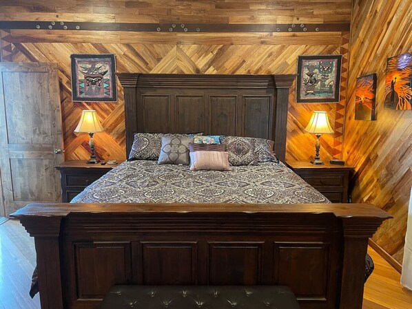 A wooden-paneled bedroom with a large bed, two bedside tables, and wall art. - Magic Skies Hawaiian Kingdom Hula Room (Hilo)