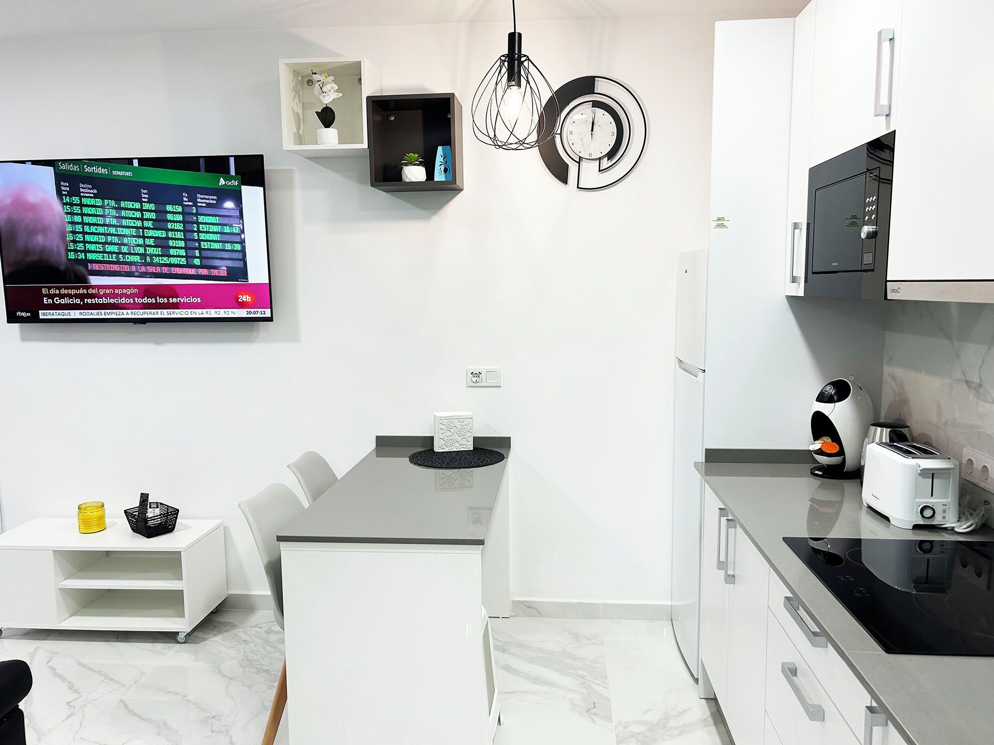 A modern kitchen with a built-in oven, a coffee machine, and a TV displaying flight information.