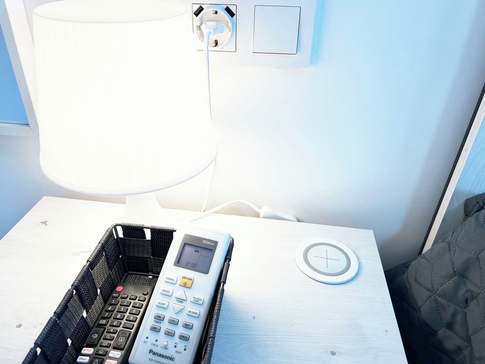 Ein Zimmer mit einem weißen Nachttisch, einer Panasonic-Klimaanlage, einer Lampe und einer Steckdose.