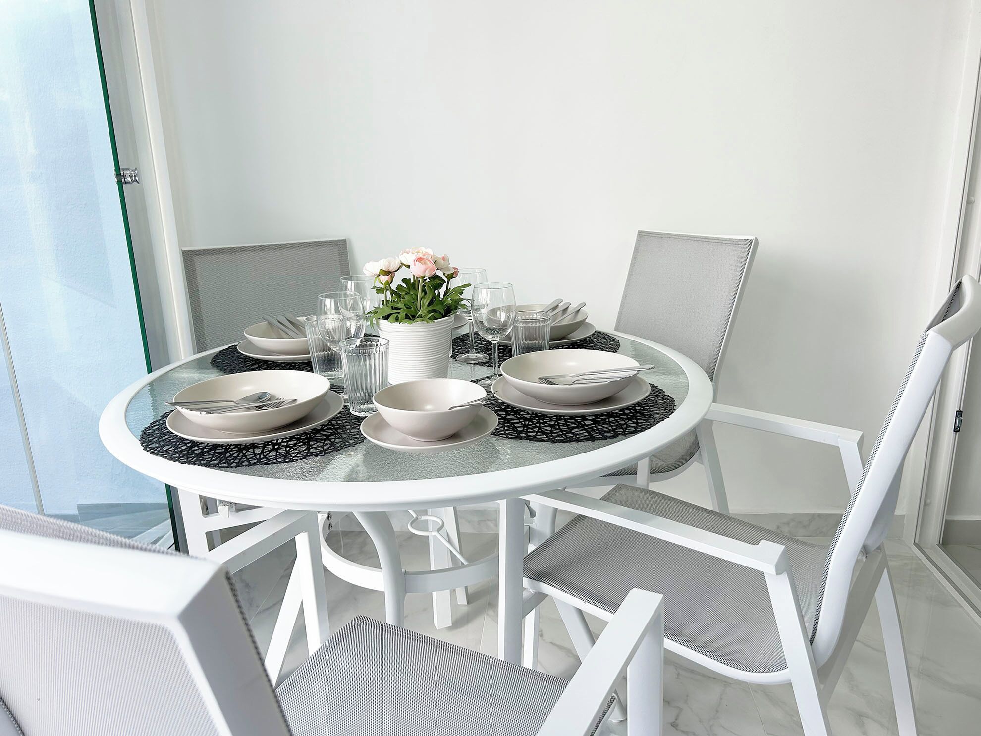 Una mesa redonda de vidrio con sillas blancas, dispuesta con platos, tazones, vasos y una planta en maceta.