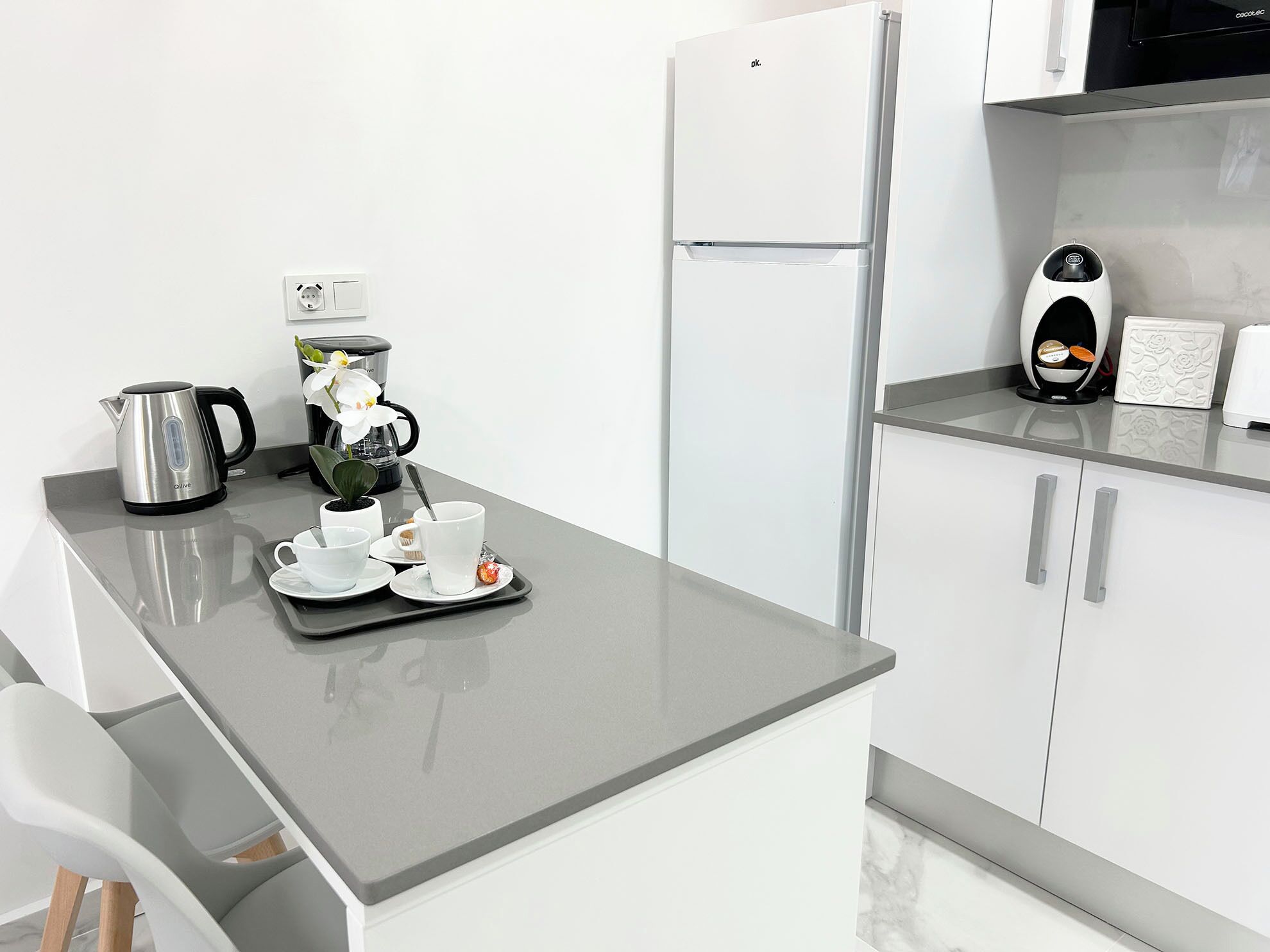 A modern kitchen with a refrigerator, coffee maker, kettle, and a tray with cups and a flower.