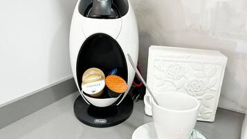 Una cafetera con dos cápsulas de café, una cuchara y una taza blanca sobre una mesa.