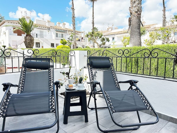 Two lounge chairs with a small table and a plant between them, set on a balcony with a view of white buildings and palm trees.