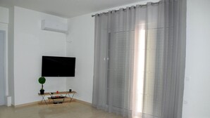 A room with a flat-screen TV mounted on the wall, a small wooden table with a plant and a remote control, and a large window with sheer curtains. - Comfort Holidays (Messini)