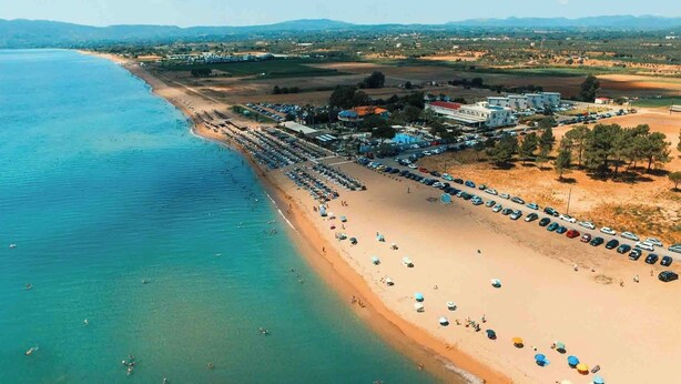An aerial view of a beach with rows of beach umbrellas and sun loungers, a parking lot, and buildings in the background. - Comfort Holidays (Messini)