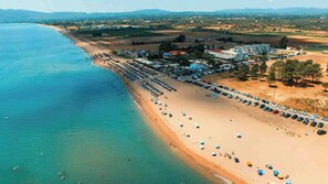 Eine Vogelperspektive zeigt einen Strand mit Reihen von Strandsonnenmatten und Sonnenschirmen, einen Parkplatz und Gebäude im Hintergrund.