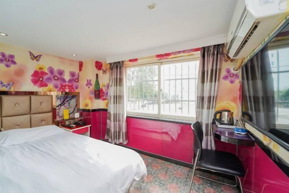 A hotel room with a bed, a desk, a chair, a window with curtains, and a wall with floral patterns.