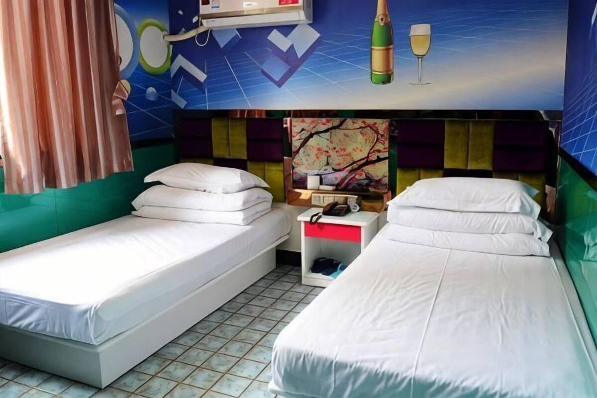 A hotel room with two single beds, a small table with a lamp, a wall-mounted shelf, and a colorful abstract painting.