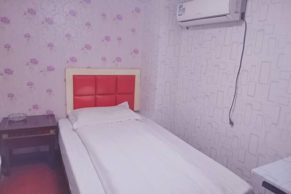 A small, neatly made room with a single bed, a nightstand, and an air conditioning unit on the wall.