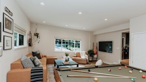 A living room with a pool table, a sofa, a television, and a window with a view of greenery.
