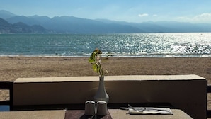 Uma mesa com um vaso de flores, saleiro e pimenteiro e talheres numa varanda com vista para uma praia e montanhas ao fundo.