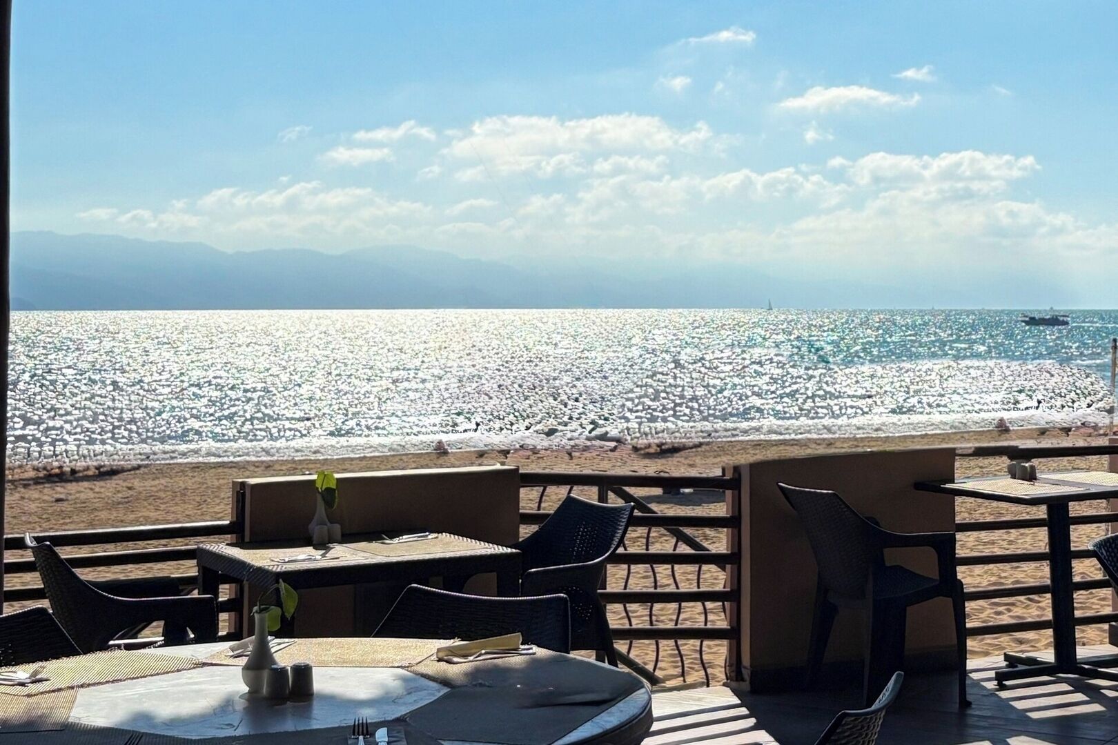 An outdoor seating area with tables and chairs overlooking a beach with a rocky shoreline and a ship in the distance.