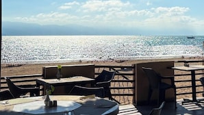 Uma área de estar externa com mesas e cadeiras com vista para uma praia com costa rochosa e um navio ao longe.