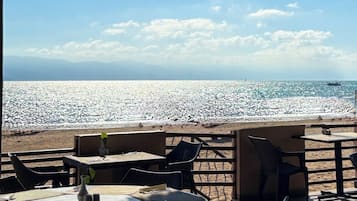 An outdoor seating area with tables and chairs overlooking a beach with a rocky shoreline and a ship in the distance.