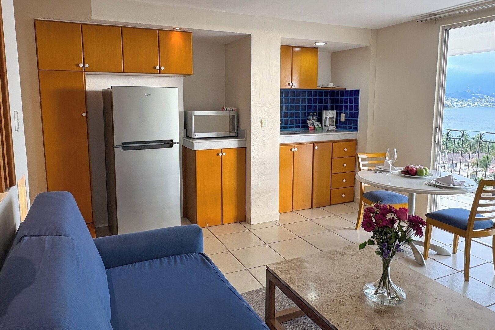 A modern kitchen with wooden cabinets, a refrigerator, a microwave, and a dining area with a table and chairs.