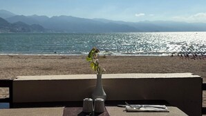 A table with a vase of flowers, salt and pepper shakers, and cutlery on a balcony overlooking a beach with mountains in the background. - Partial ocean view, exclusive amenities! (Puerto Vallarta)