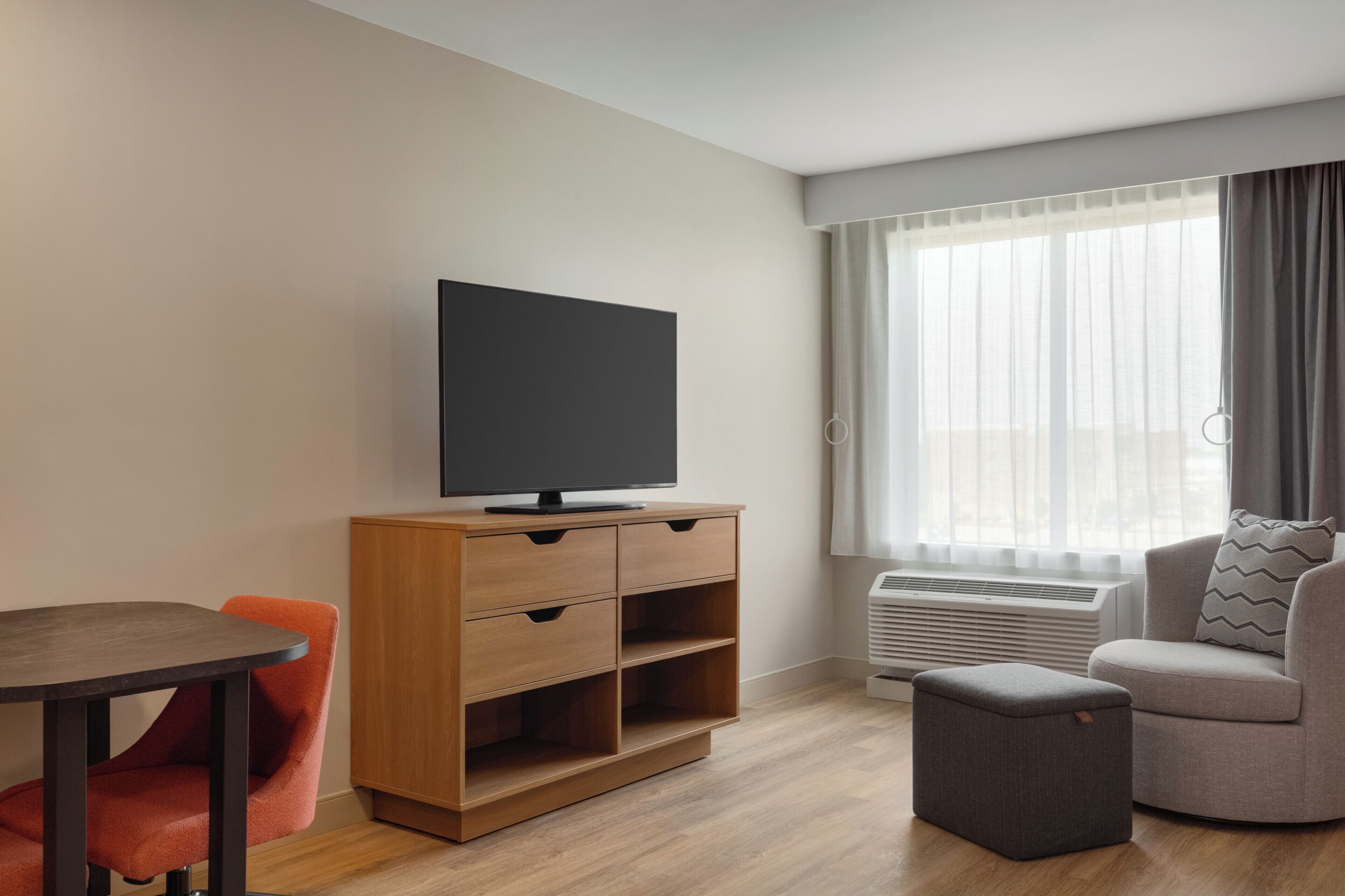 A modern living room with a flat-screen TV on a wooden cabinet, a grey sofa, a small ottoman, and a dining table with orange chairs.
