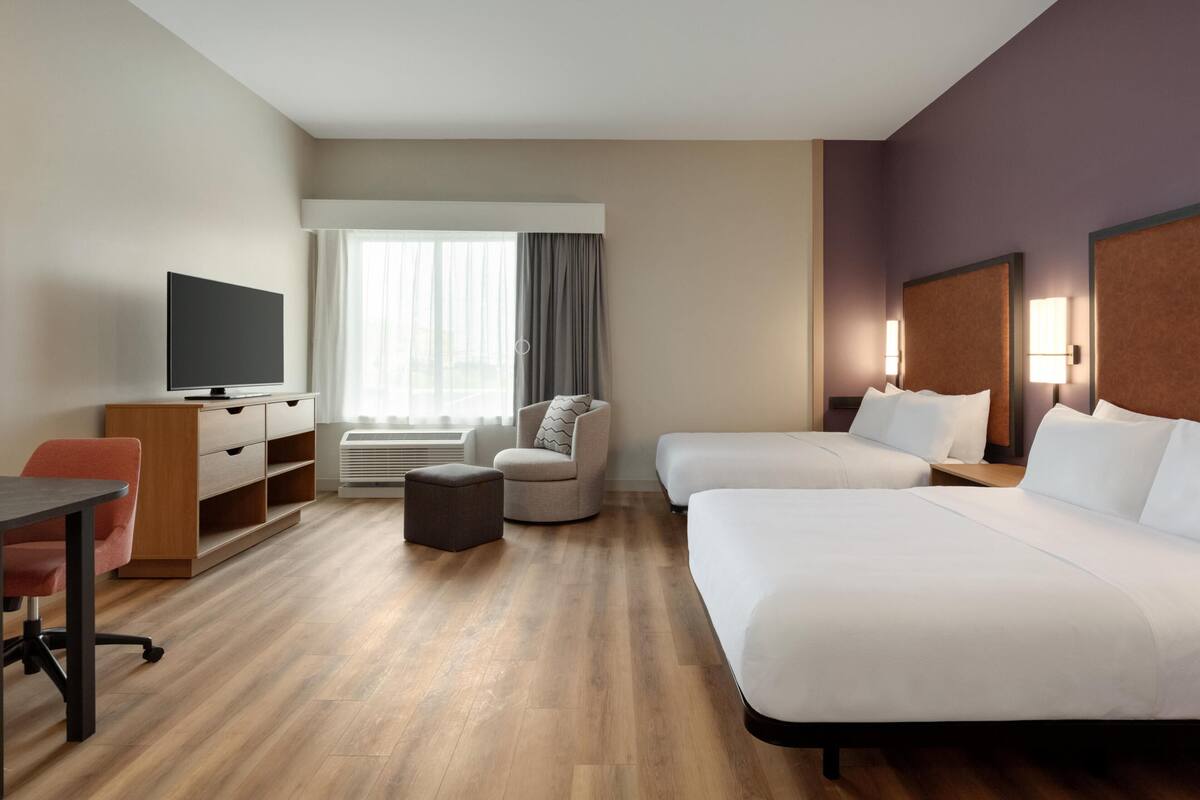 Een hotelkamer met een bed, een bureau, een stoel, een televisie, een raam met gordijnen en een nachtkastje met een lamp.