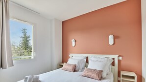 A neatly made bed with white linens and striped pillows, a window with curtains, and a wall-mounted light fixture. - Maisonnette Confortable | Terrace Privée + Parking (Aigues Mortes)
