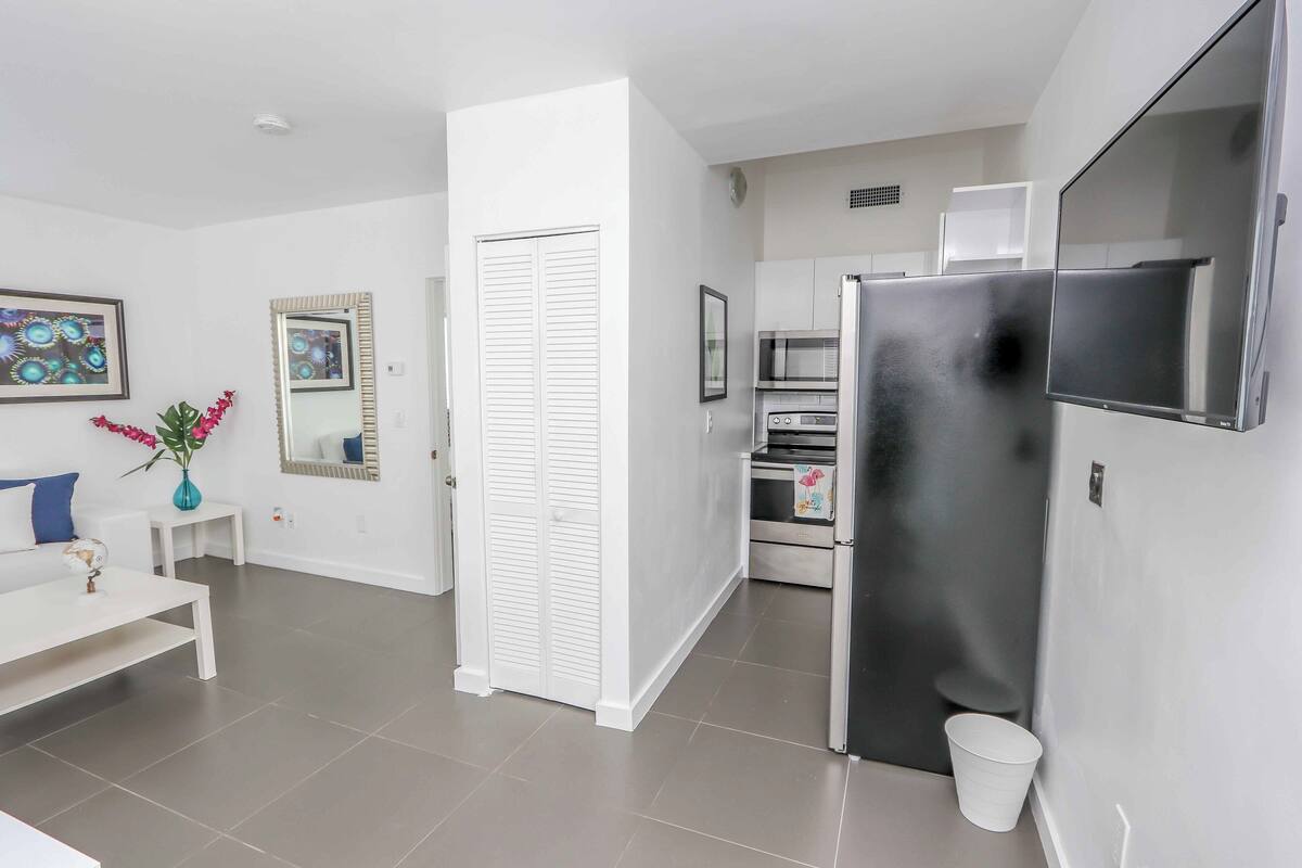 A modern kitchen with a refrigerator, oven, and microwave. There is a white door and a small white table with a vase of flowers.