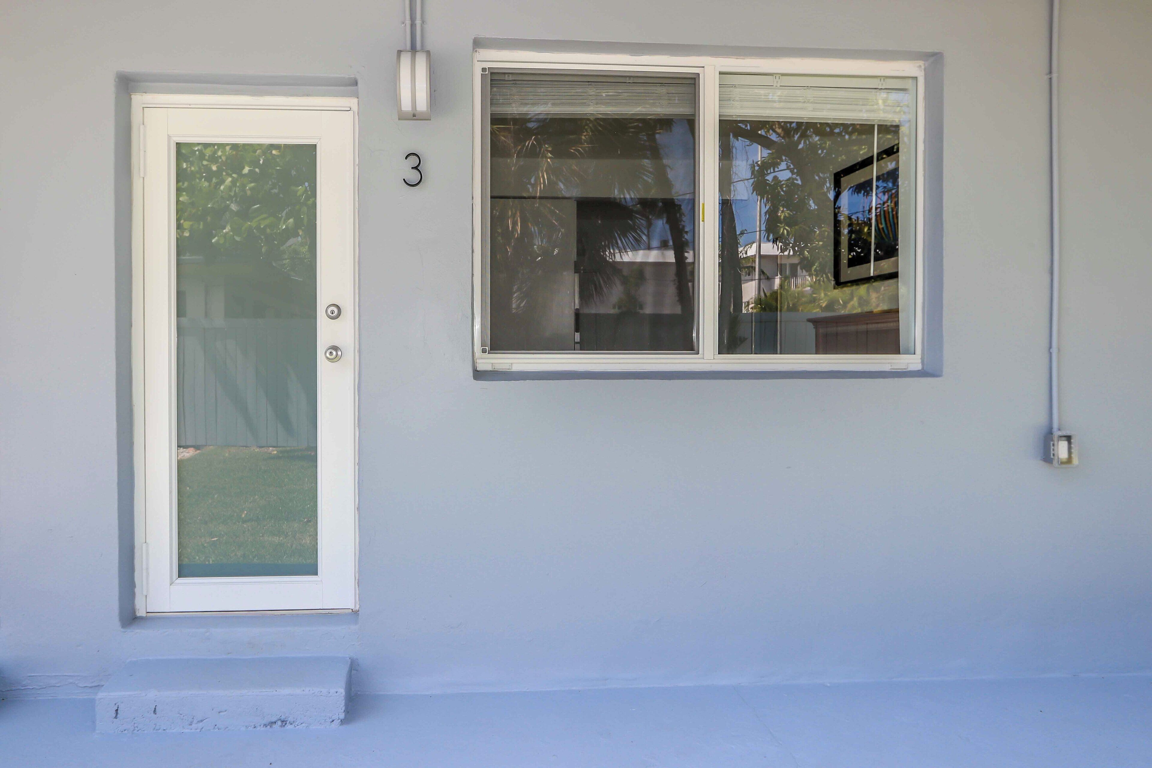 Un numéro de maison, le chiffre « 3 », peint en blanc sur un mur bleu clair, une porte blanche avec un panneau de verre et une fenêtre avec un cadre blanc.