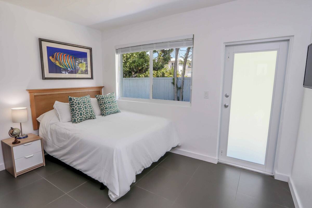 A bedroom with a bed, a nightstand, a framed picture, a window with a view, and a door.