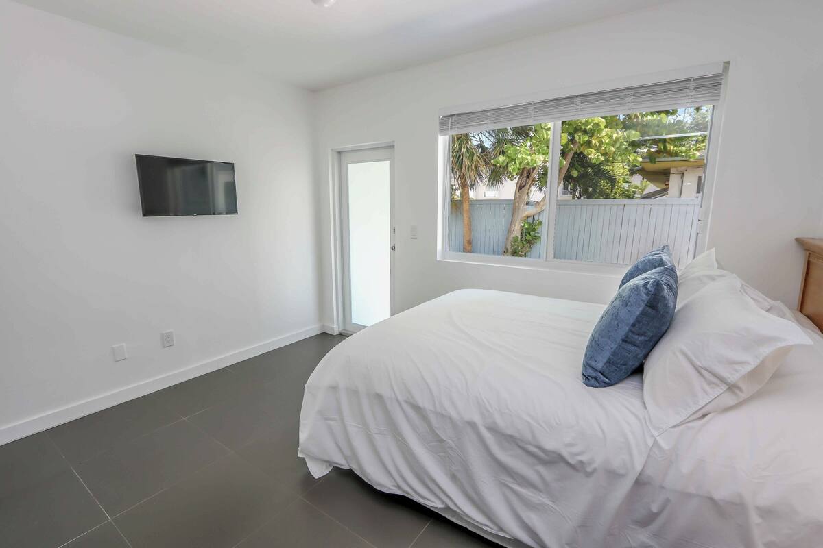 A bedroom with a bed, a TV mounted on the wall, and a window with a view of trees and a fence.