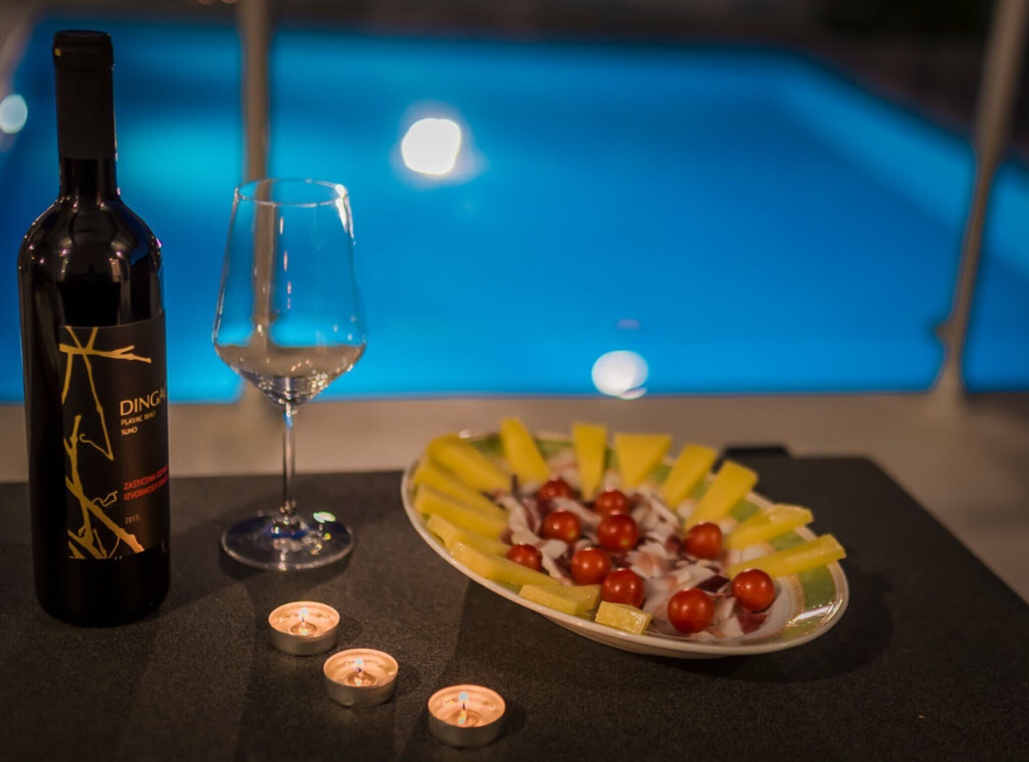 A bottle of wine, a glass, and a fruit platter on a table with candles, near a pool.