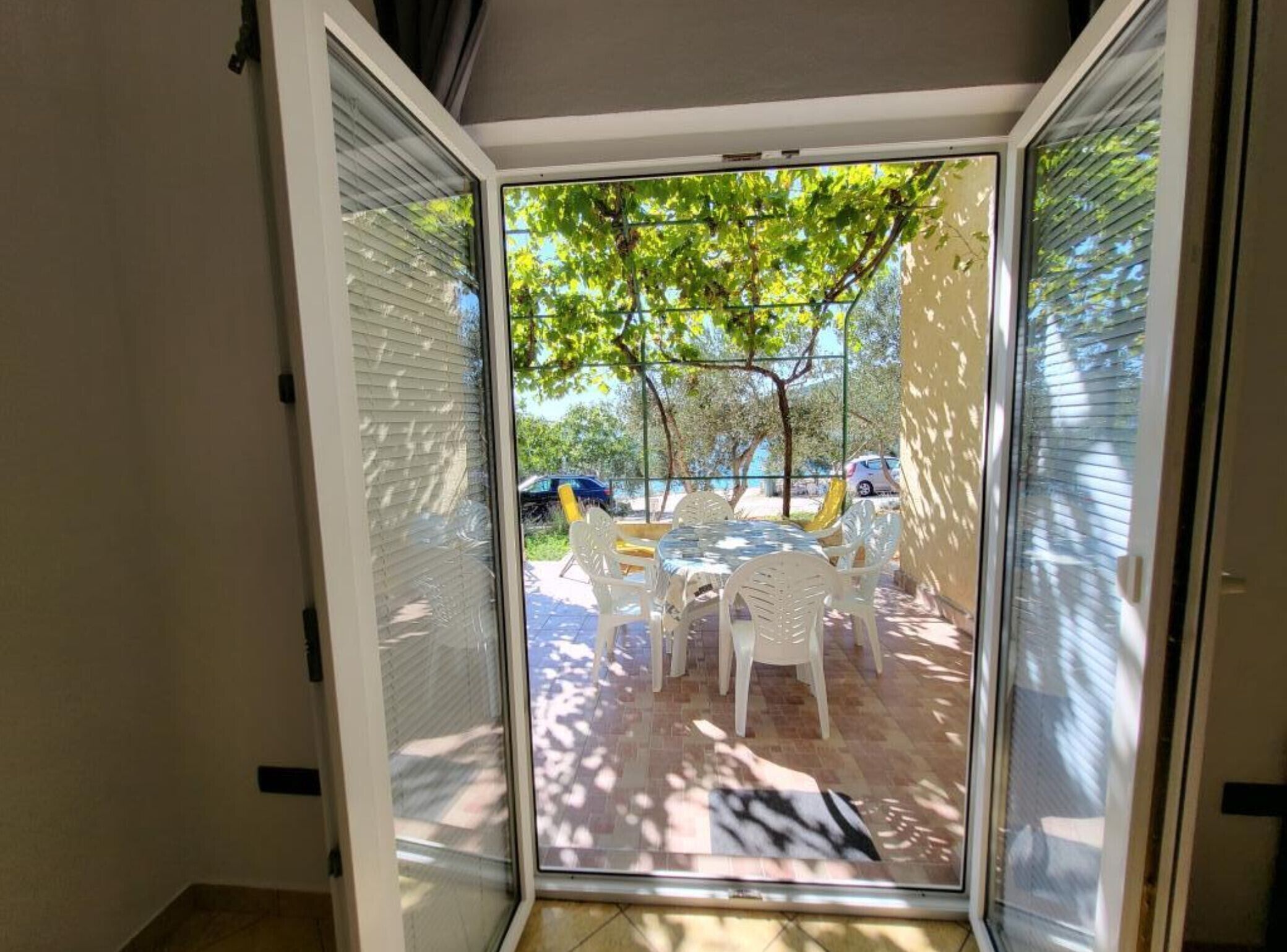 Une porte-fenêtre coulissante donne sur un patio extérieur aménagé avec une table et des chaises, entouré d’arbres et offrant une vue sur les voitures garées.