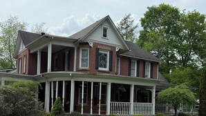 Une maison à deux étages avec une véranda qui fait le tour de la maison, une façade en briques et une pelouse bien entretenue.