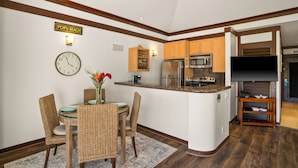 A kitchen with wooden cabinets, a microwave, and a refrigerator. There is a dining area with wicker chairs and a table. - New Renovated April 2025 Charming Apartment In Delightful Kiahuna with AC, (Koloa)