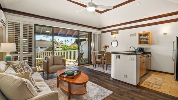 A spacious living area with a sofa, coffee table, and a dining set. A kitchen area is visible with a refrigerator and a countertop. - New Renovated April 2025 Charming Apartment In Delightful Kiahuna with AC, (Koloa)