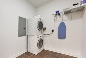 A laundry room with stacked washers and dryers, a blue ironing board, and a shelf with various items.
