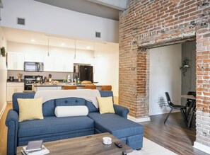 Una sala moderna con un sofá seccional, un comedor con pared de ladrillo y una cocina al fondo.