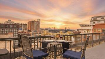 Varanda com duas cadeiras e uma mesa, com vista para a paisagem urbana com edifícios e uma ponte ao pôr do sol.