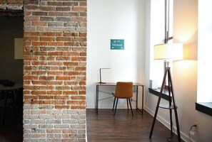 A room with a brick wall, a desk with a chair, a standing lamp, and a sign indicating the direction to Pittsburgh.