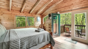 A wooden cabin bedroom with a bed, a chair, and a balcony with a view of the forest.