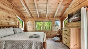 A wooden cabin bedroom with a bed, dresser, and a window with a view of greenery.