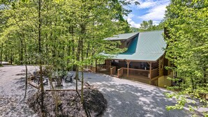 A lodge with a green roof, surrounded by trees, featuring a covered deck and a gravel driveway.