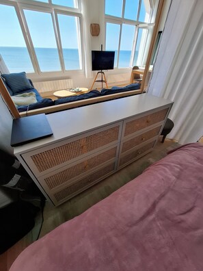Una habitación con una cama, un escritorio con una computadora portátil, un televisor sobre un pedestal y vistas al mar a través de amplios ventanales.