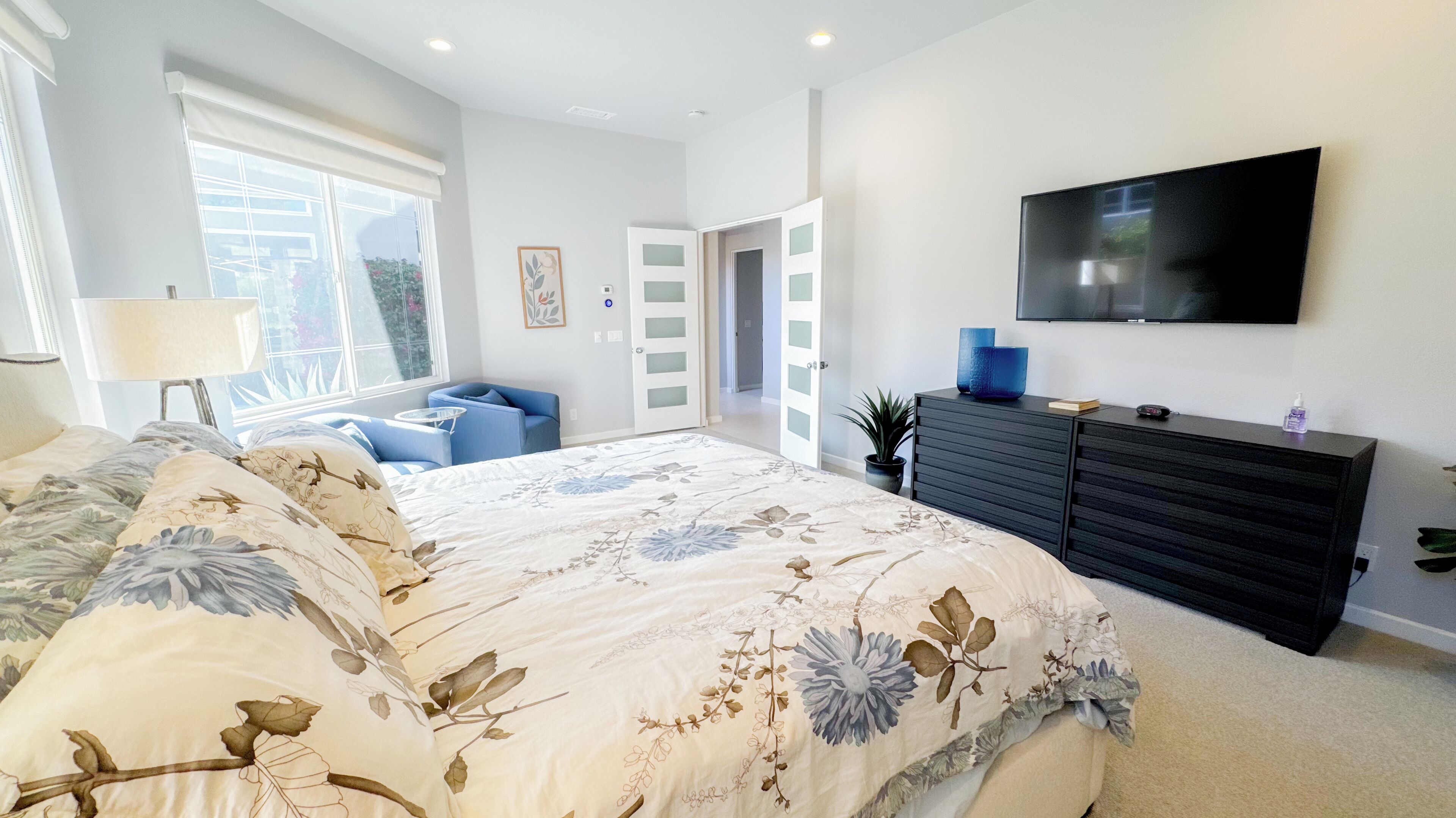 A bedroom with a large bed, a TV mounted on the wall, a dresser, and a window with a view.
