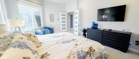 A bedroom with a large bed, a TV mounted on the wall, a dresser, and a window with a view.