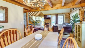 Interior acogedor de una cabaña de troncos con comedor, sillas de madera, una araña y una sala de estar con chimenea.