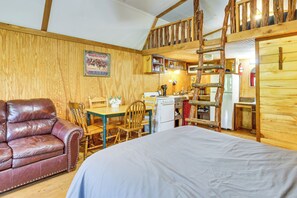A rustic cabin interior with a wooden dining table, a sofa, a bed, a kitchen area, and a loft space.