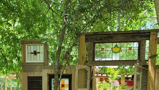 Una estructura rústica de madera con un ventanal de vitral y una puerta de madera, rodeada de abundante vegetación y un camino de piedra.
