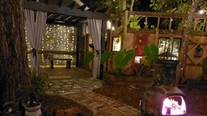 Un espacio al aire libre cubierto con un sendero de piedra, un banco de madera, plantas en macetas y un elemento decorativo de fuego.