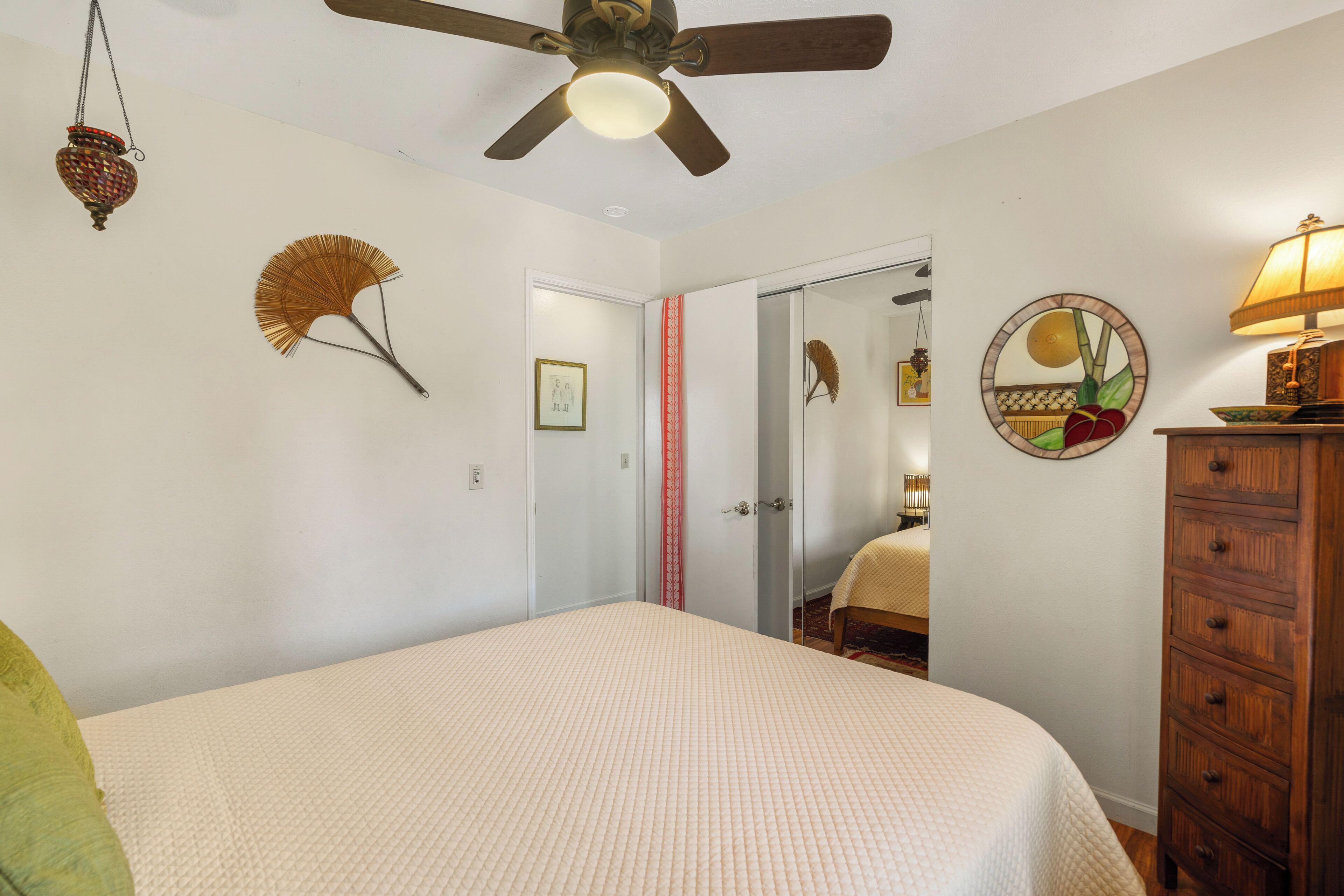 Une chambre à coucher comprenant un lit, un ventilateur de plafond, une commode, un miroir et une porte donnant sur une autre pièce.